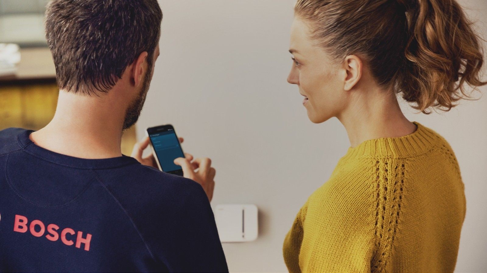 Person using a smartphone while another stands nearby, with a wall-mounted device in the background.